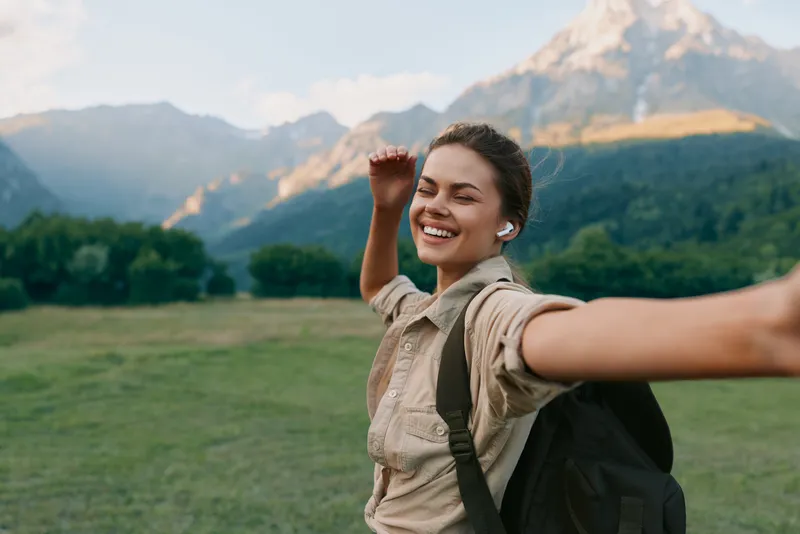 Smiling woman traveler with backpack posing outdoors in a green valley, mountains in the background, depicting an active lifestyle and joyful exploration for social sharing