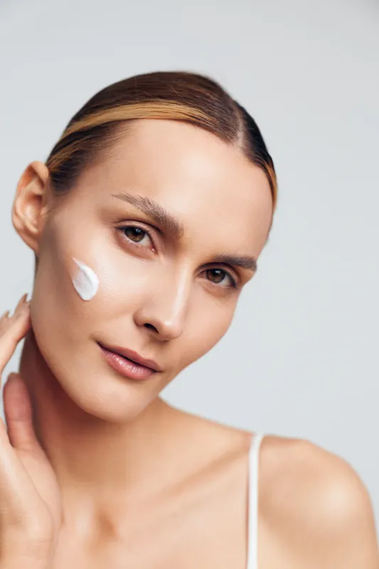 Smiling woman with smooth skin applying white cream to her cheek. Close up on a light background. Concept of skincare, self care, and wellness. Soft studio lighting creates a calm, fresh mood.
