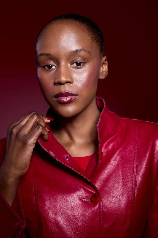 Confident black woman with dark skin wearing a red leather jacket poses against a red background. Studio portrait captures elegance, fashion, and beauty with glossy makeup and smooth skin.
