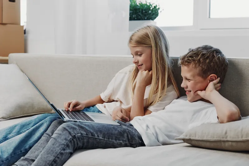 Two children relax on a sofa using a laptop together in a bright living room. The girl and boy enjoy technology and leisure time, illustrating family bonding and digital learning.