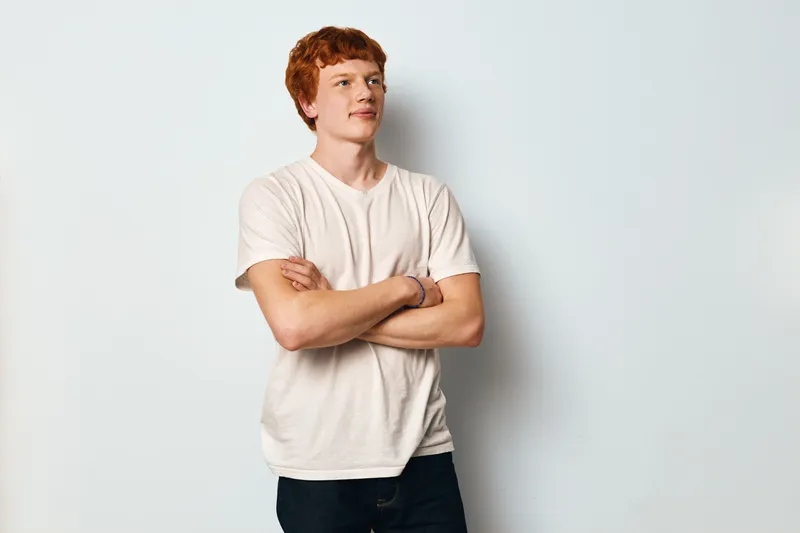 Young man with red hair standing with arms crossed looking confident in casual white t shirt and jeans on plain light background.
