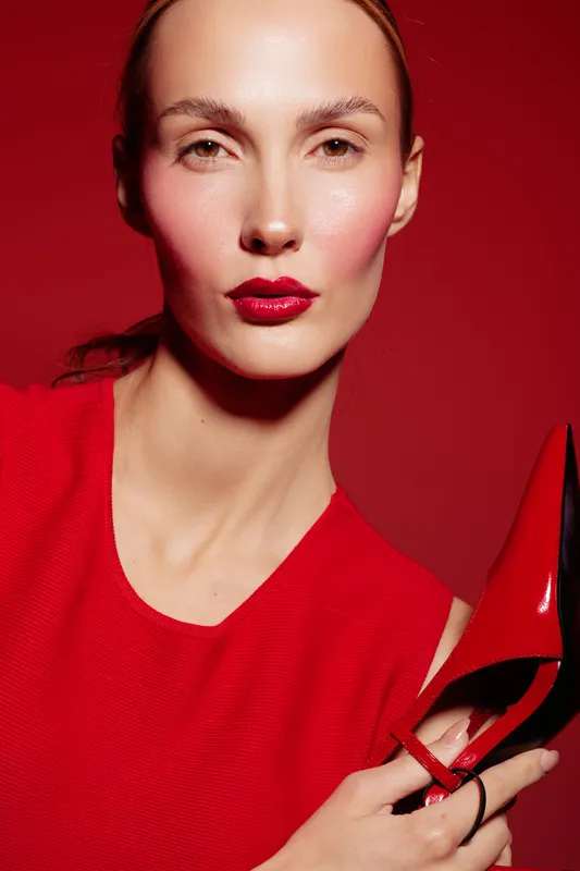 Close-up of a woman with red lipstick and red dress holding a red high heel shoe against a red background. Concept of fashion, beauty, and style with monochrome tones.