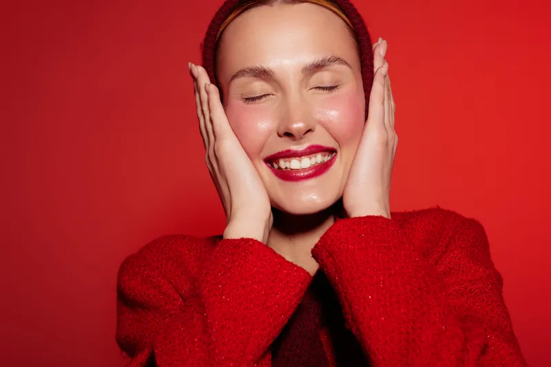 Smiling woman with closed eyes and red lipstick touches her face with hands. She wears a red sweater and headband on red background. Concept of happiness, beauty, and skincare.
