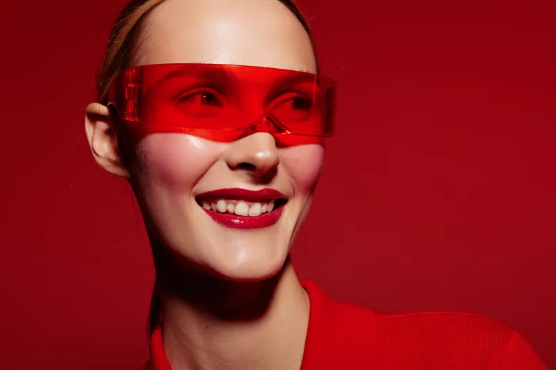 Smiling woman wearing red transparent glasses and red outfit poses on red background. Close-up portrait with bright makeup and glossy lips. Concept of fashion, style, and modern accessories.