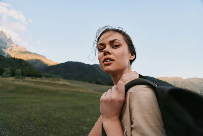 Young woman with a backpack glances back on a sunlit mountain trail, ready for adventure in a wide open natural landscape