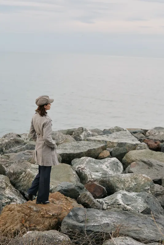 Woman in beige coat and hat stands on rocky shore looking at calm sea under cloudy sky. Concept for solitude, contemplation, nature and autumn season outdoors.