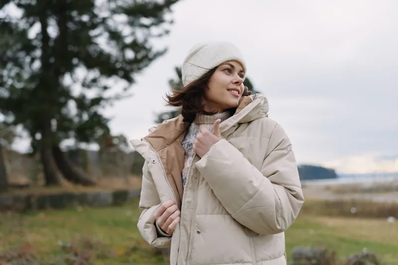 Woman in a beige winter coat and knit beanie stands in an open field, smiling softly as she adjusts the collar. A cozy outdoor fashion portrait with calm mood and natural winter light.