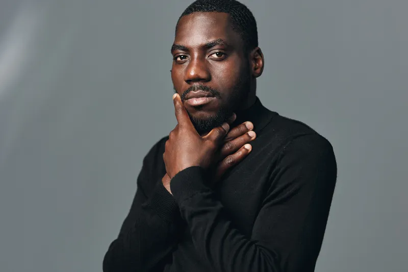 African American man with dark skin and short hair in creative visual designer style posing with hand on chin and neck against gray background in studio portrait.