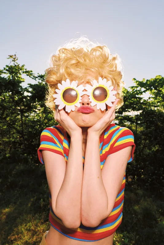 Playful woman with curly blonde wig and sunflower sunglasses poses outdoors wearing a striped crop top, radiating retro film color and a carefree lifestyle vibe