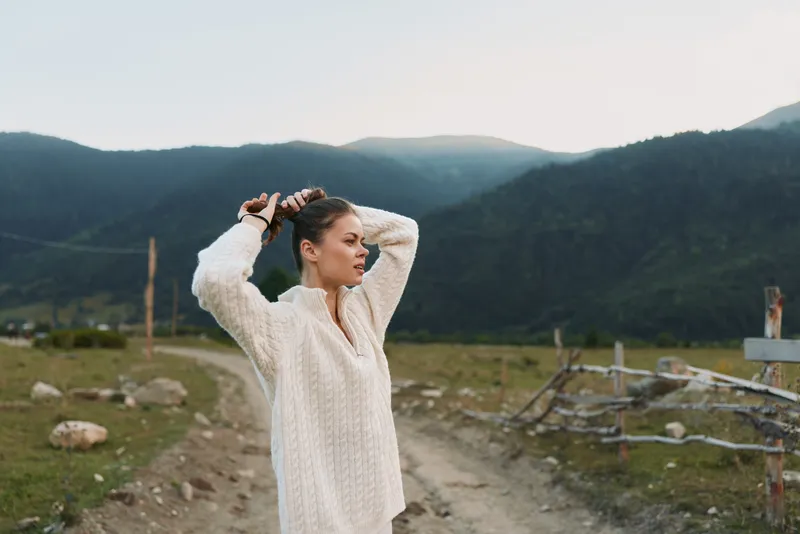 A woman in a light sweater stands on a dirt road in a rural valley, adjusting her hair. Scenic mountains frame the scene, creating a calm lifestyle moment.