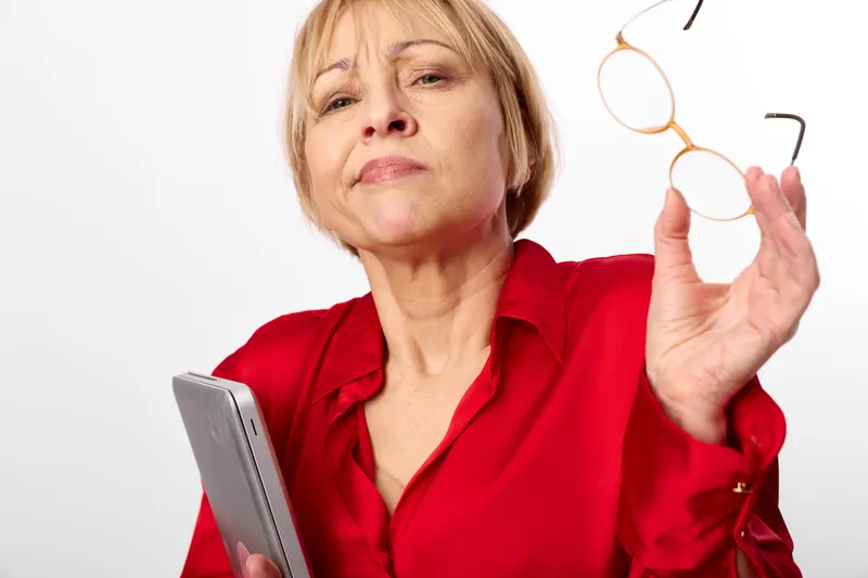 Senior woman holding glasses by tablet, adjusting eyewear, soft studio on white background, vision care concept, technology for health, calm expression