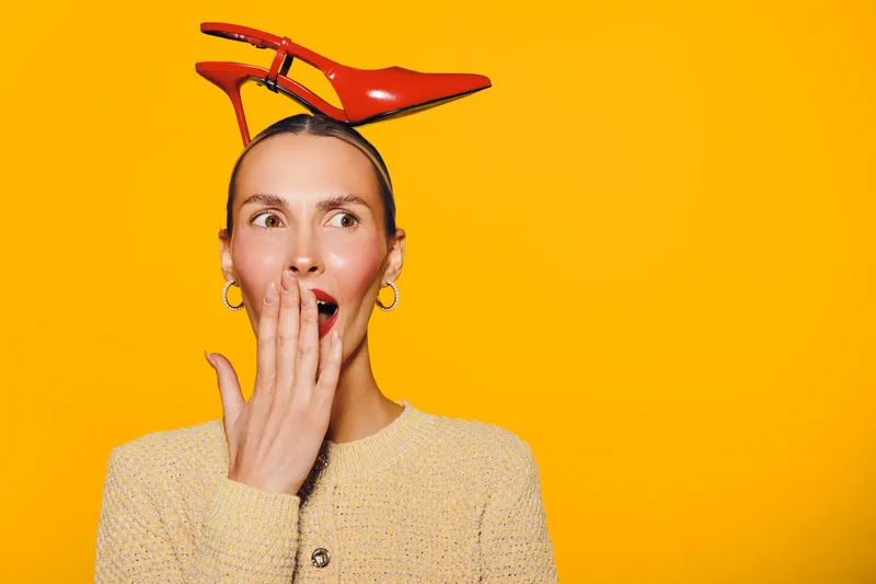 Surprised woman with red high heel shoe on head covers mouth with hand. Bright yellow background and casual beige sweater create playful and quirky fashion concept with unexpected style.