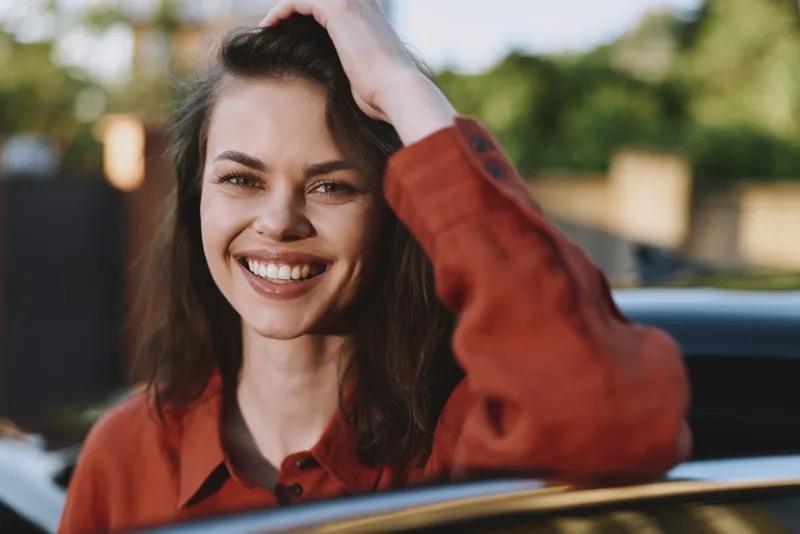 Smiling brunette young woman leans on a car window outdoors, enjoying warm sunlight, a relaxed carefree vibe, and a casual everyday moment with gentle breeze and soft background greenery