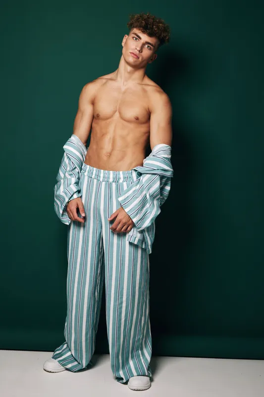 Young man with muscular body posing shirtless wearing striped loose pants and open shirt against dark green background in studio for fashion and fitness concept.
