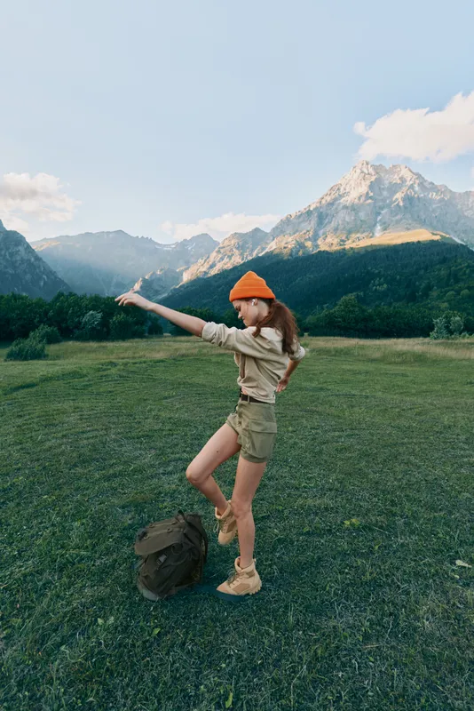 Young woman in casual outfit standing on green grass field with backpack pointing at mountain peak under blue sky with clouds, outdoor lifestyle adventure concept