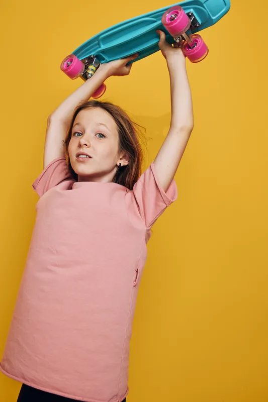 Young girl in a pink t-shirt holds a turquoise skateboard above her head against a yellow background. Studio portrait showing playful and confident attitude.