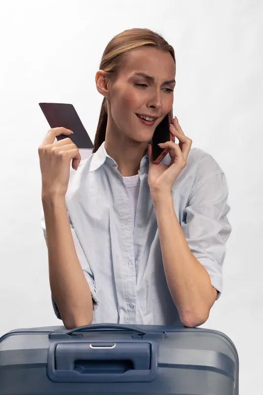 Young woman holds passport and talks on phone while standing behind suitcase, preparing for travel. Concept of trip planning, communication, and vacation readiness.