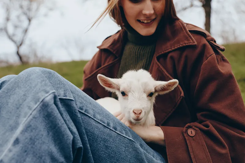 Young woman in a brown coat holds a white baby goat while sitting outdoors on grass during a cloudy day, enjoying a calm and intimate moment with the animal.