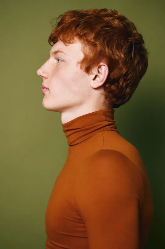 Profile of young man with red hair wearing brown turtleneck sweater against green background with side view portrait and neutral expression in studio lighting.