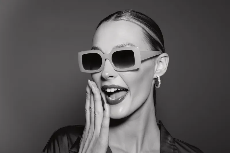 Stylish white woman with light skin wearing large square sunglasses and hoop earrings poses playfully in black and white studio portrait.