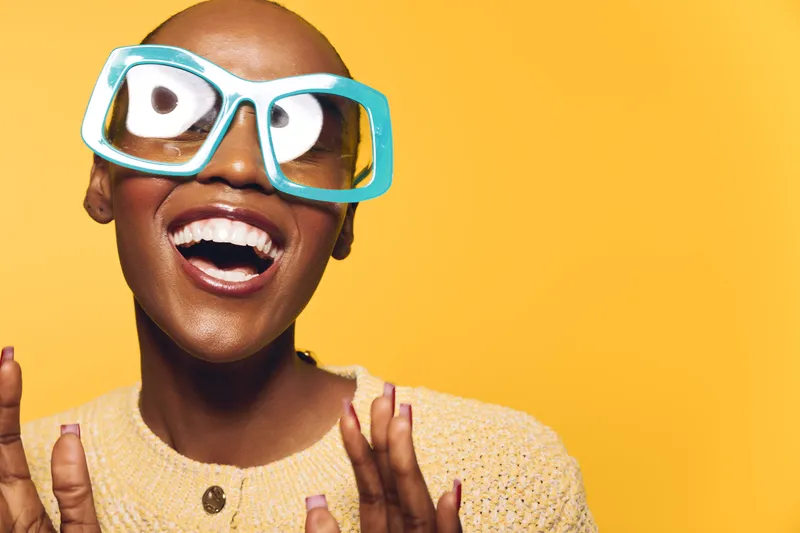 Happy African American woman with dark skin wearing large turquoise glasses and yellow sweater smiling widely on bright yellow background. Concept for joy, fashion, and self-expression.