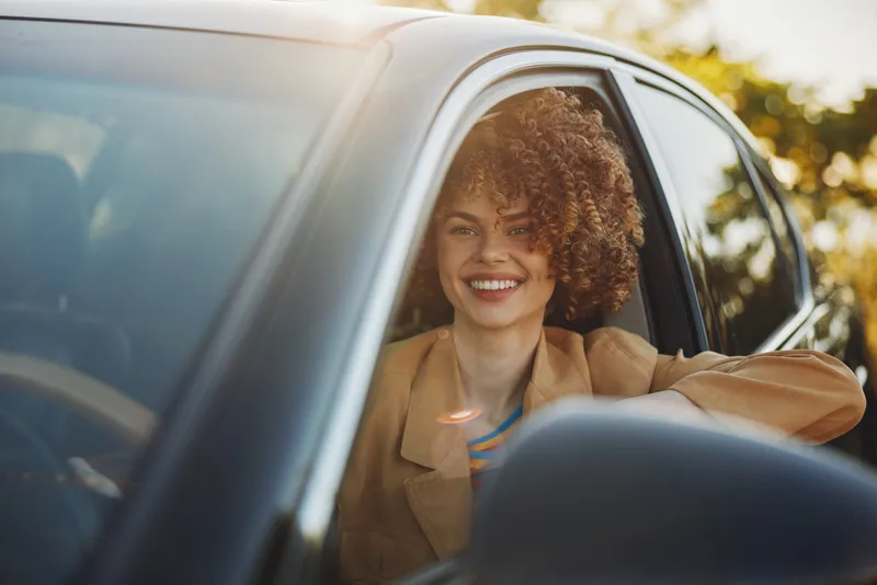 A smiling woman driver in a car poses for a lifestyle portrait with outdoors scenery and warm retro film tones, conveying casual confidence, happiness, and relaxed travel.
