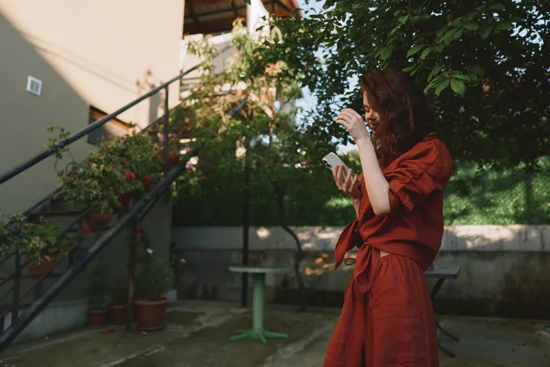 Young woman in a red dress stands in a sunlit courtyard, adjusting her hair with a carefree, confident smile, conveying casual lifestyle vibes and fashion portrait potential