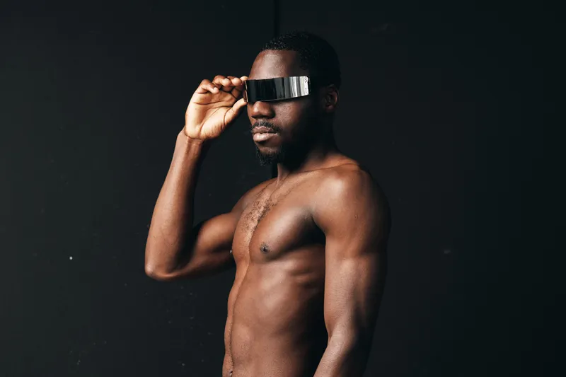 Creative conceptual portrait of shirtless African man wearing futuristic visor glasses in visual style with dark background and strong lighting for modern fashion.