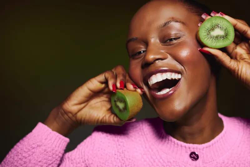 Happy dark skinned African woman with smooth skin holds fresh kiwi halves near face. Close-up portrait with natural makeup and joyful smile on dark green background. Concept of healthy food.