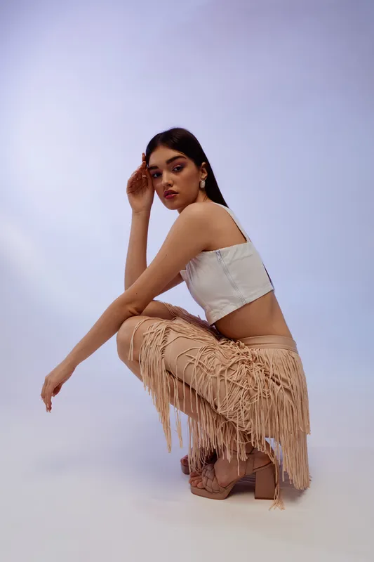 Young woman in a white crop top and beige fringed skirt poses in a studio with soft lighting. Concept of woman beauty fashion and modern style with confident expression.