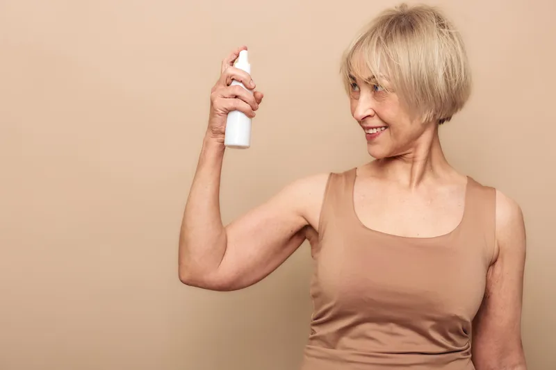 Smiling mature woman with short blonde hair sprays facial mist on neutral beige background. Concept of skincare routine, hydration, self-care, and natural beauty for older adults in studio lighting.