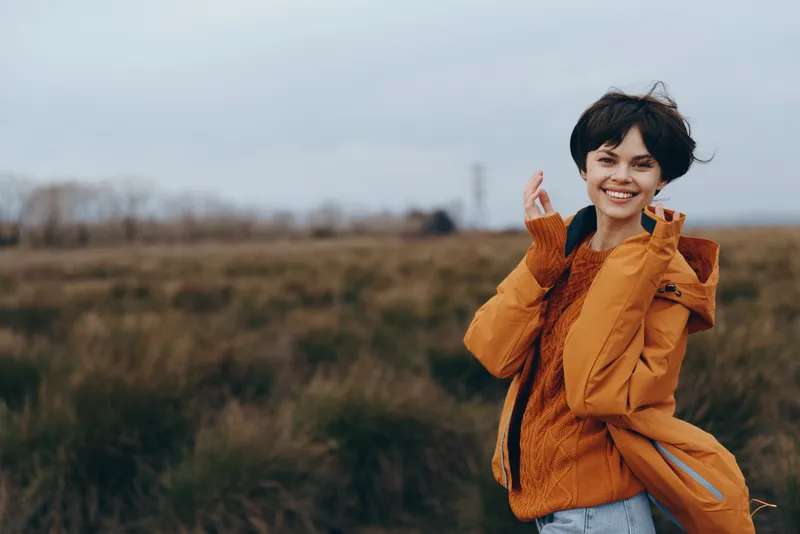 Woman lifestyle outdoors portrait, autumn field, orange jacket, smiling with relaxed pose, hand near hair, soft daylight, blurred grass and trees background, countryside calm atmosphere