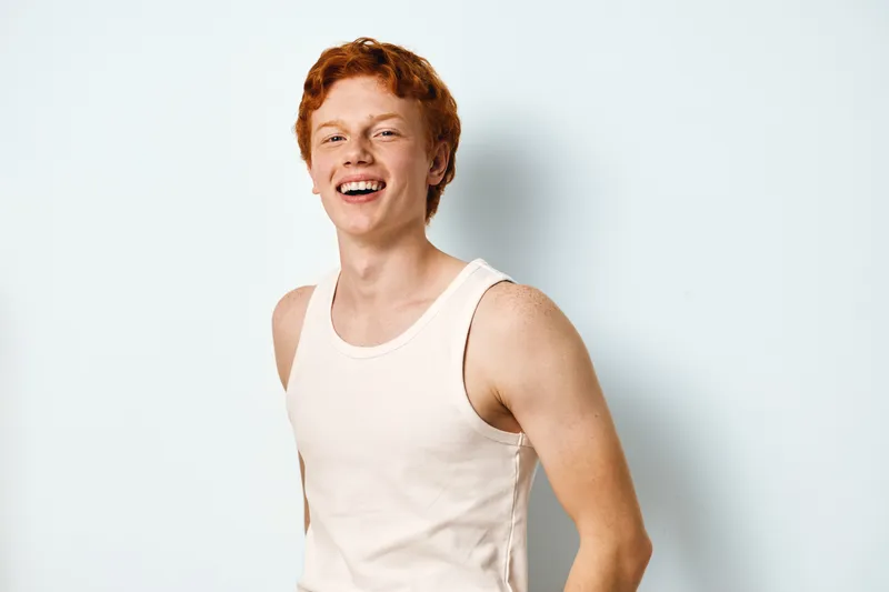 Young man with red hair and freckles wearing white sleeveless shirt smiling and laughing against white background. Casual, happy, portrait and positive expression on face.