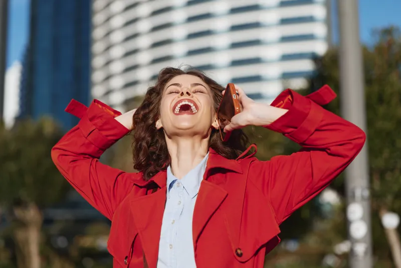 Young woman in a red coat laughs joyfully while holding a phone to her ear outdoors in an urban environment on a sunny day.