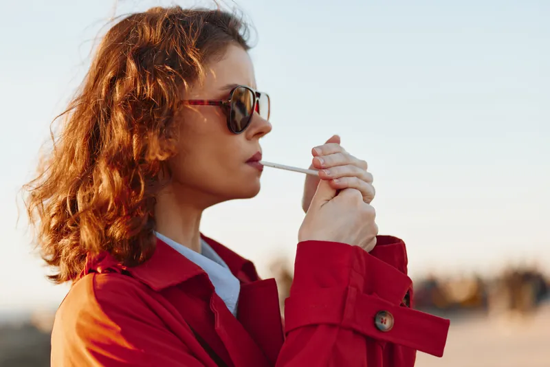 A young woman in a red coat and sunglasses lights a cigarette outdoors during sunset, showing a calm and focused expression in natural light.