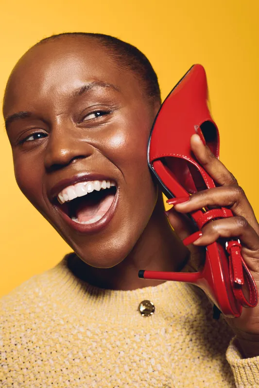 Happy African woman with dark skin holds red high heel shoe near ear, smiling widely. Yellow background and cozy sweater create a bright, joyful fashion concept.