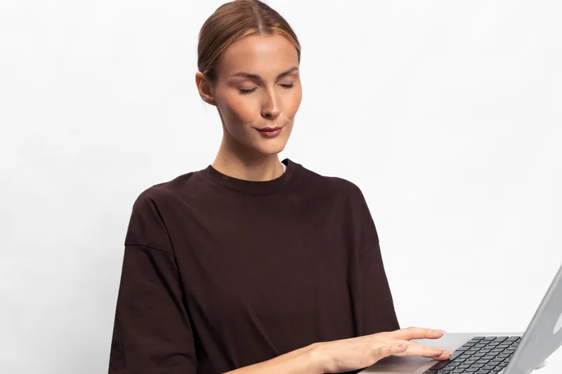 Young woman in dark sweater typing on laptop with focused expression. White background and soft lighting create professional and calm atmosphere. Concept for remote work and technology.