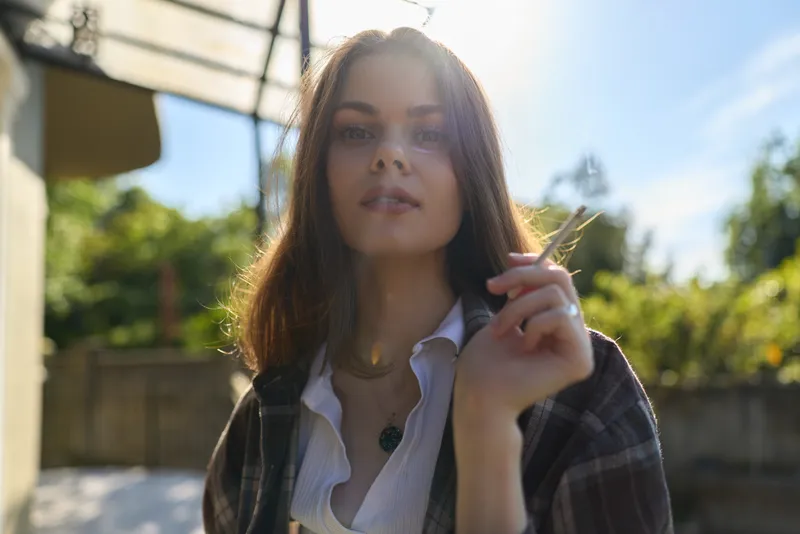 Smiling woman outdoors in daylight wearing a casual plaid shirt, holding a blade of grass. Close-up portrait with a sunlit background, showcasing relaxed lifestyle and natural beauty
