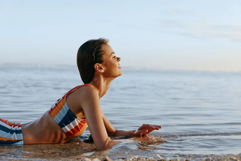 Young woman in a colorful striped swimsuit relaxes lying on her stomach at the edge of calm sea water during a peaceful sunset moment on the beach.