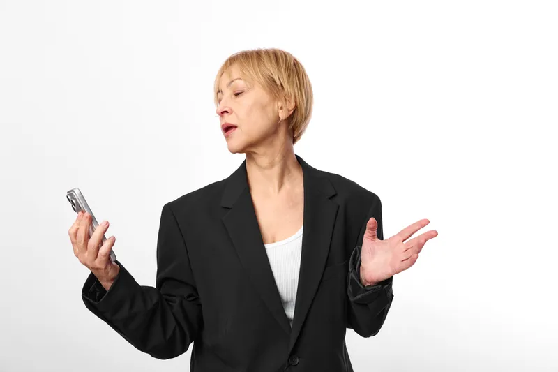 Confident businesswoman in black blazer holding smartphone and gesturing with hands, speaking in studio against white background, clean modern look, focused