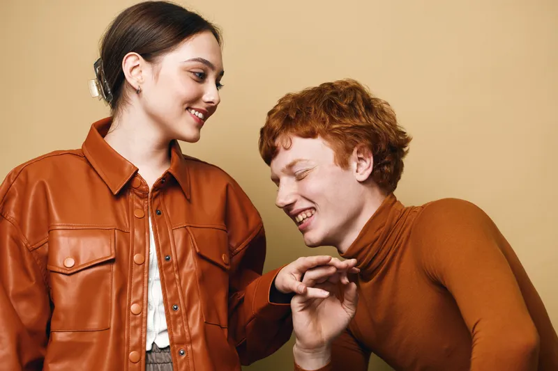 Young woman and man smiling and laughing interacting in casual autumn clothing with warm tones on neutral background. Friendship, happiness and fashion style for portrait concept.