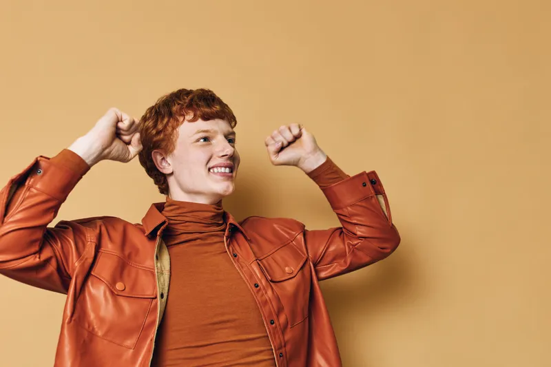 Young man with red hair wearing brown leather jacket and turtleneck celebrating with raised fists and smiling against beige background in studio. Cheerful emotional expression.