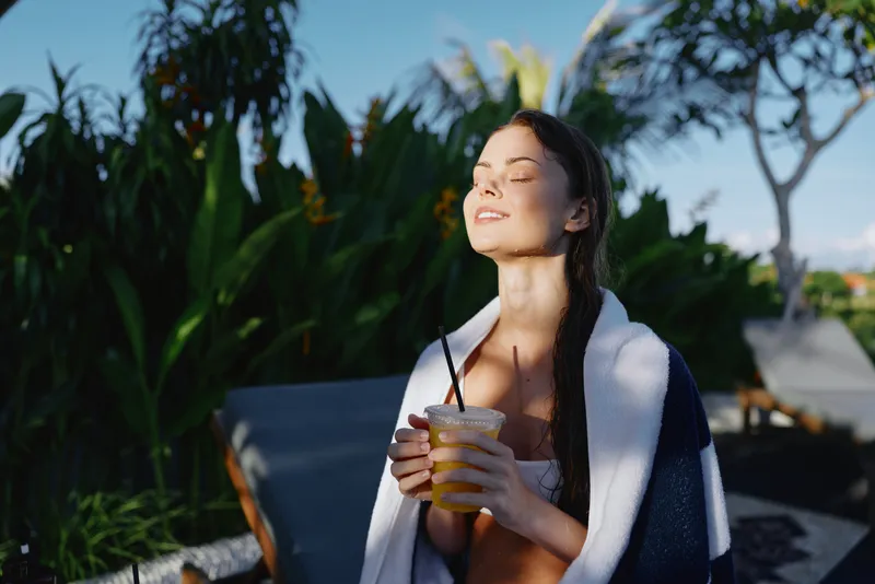 Smiling woman with towel over shoulders enjoying a cold drink outdoors, basking in sunlight. A leisure and wellness lifestyle moment captured in warm, sunlit scenery.