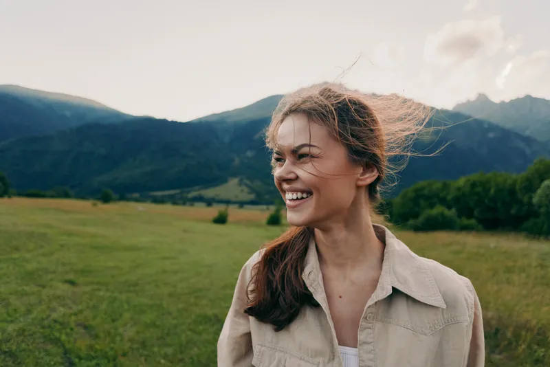 Smiling woman in casual shirt outdoors, bright mountains in the distance, open field under daytime sun, lifestyle portrait conveying happiness, freedom, and connection with nature.