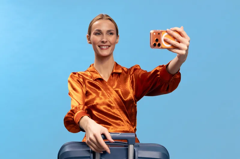 Young woman in orange blouse takes a selfie with a smartphone while holding a suitcase handle on blue background. Concept of travel, vacation, and modern lifestyle with positive mood.