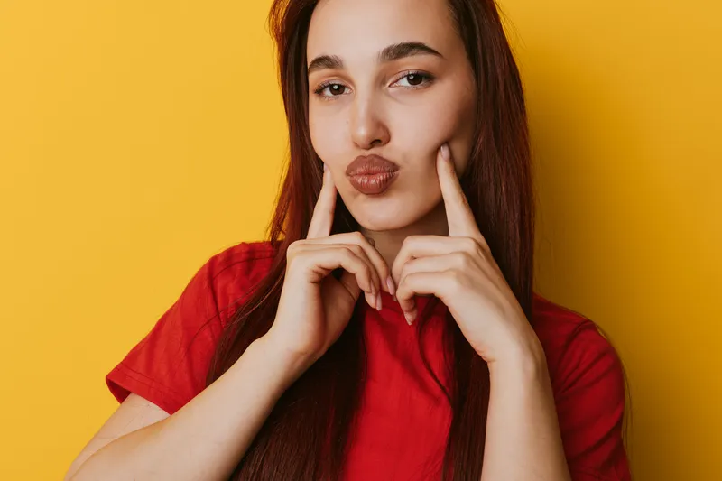 Young woman with long hair wearing red shirt posing with fingers on cheeks and lips puckered against yellow background. Portrait of playful female showing fun and confident expression.