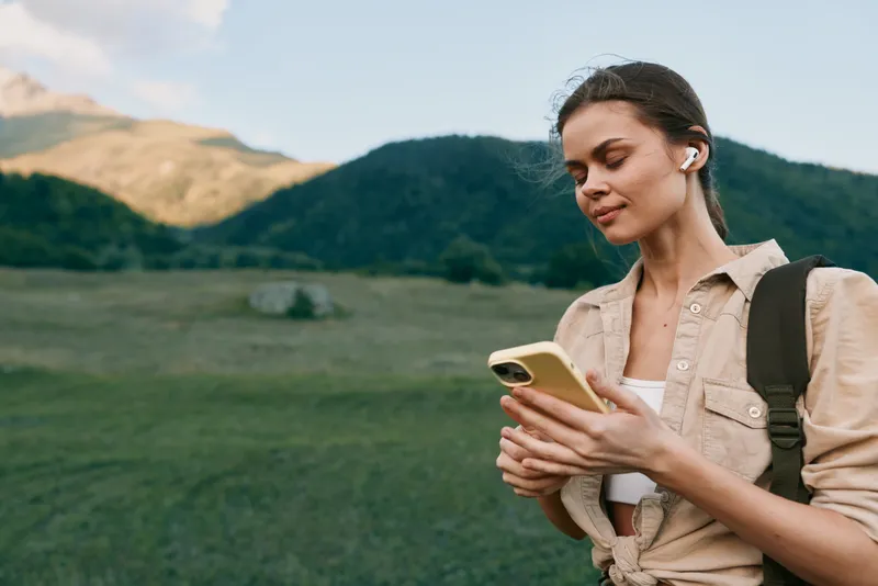 Young woman with a backpack checks her phone in a sunlit meadow, mountains in the background, calm mood, relaxed posture, outdoor lifestyle moment, gentle breeze and natural colors