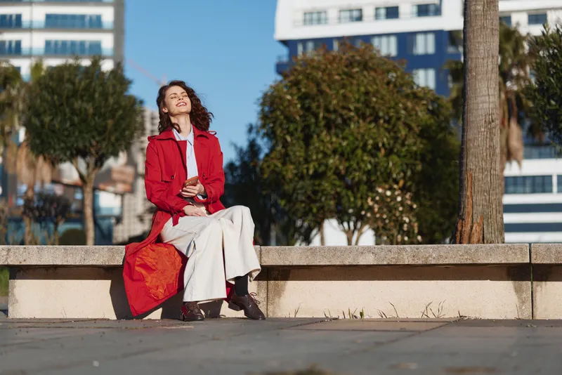 A woman sits on a concrete ledge in a red jacket and white dress, with a bright bag beside her, in an urban outdoor setting. A candid moment of casual street fashion and sunny mood.
