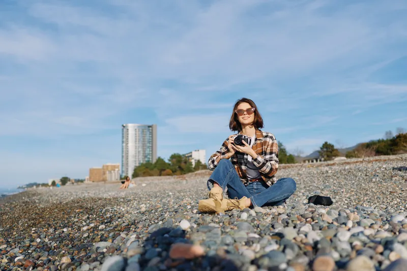 Woman sitting on a pebble beach under a clear sky, wearing sunglasses and a plaid shirt, relaxing with nearby buildings and shoreline in view.