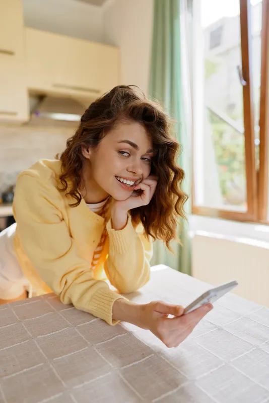 A young woman in a yellow sweater relaxes at a kitchen table, looking at her phone with a warm smile, creating a cozy home moment and casual digital connection.